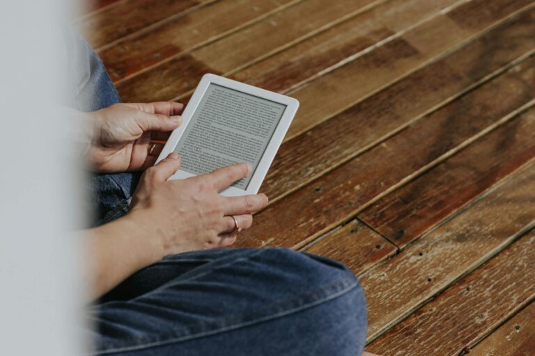 Close-up of hands holding an e-reader on a wooden floor, depicting a casual reading moment.