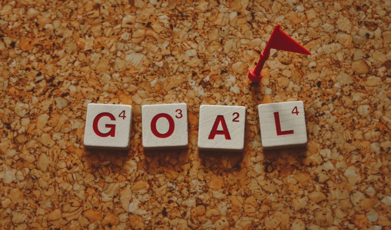 Scrabble tiles forming the word 'goal' with a small red flag on a cork board background.
