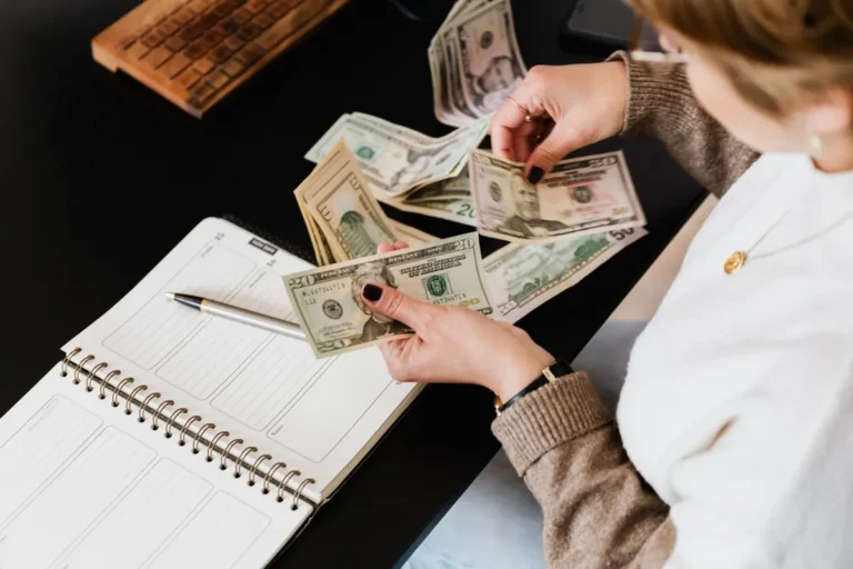 Woman organizing finances, counting dollar bills at desk with open notebook.