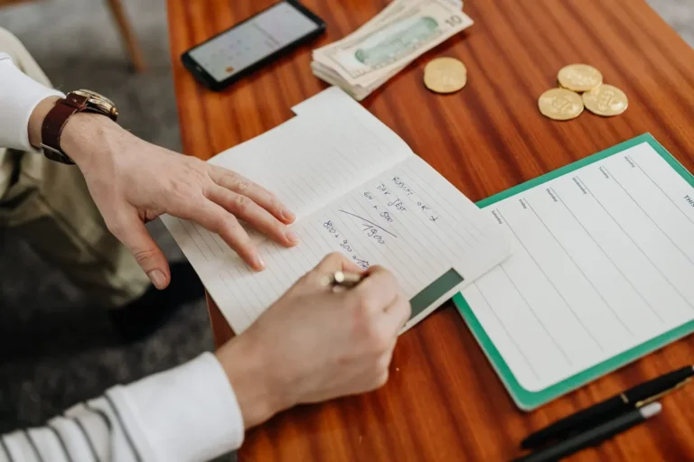 Hands writing financial calculations on notebook with money and coins on table.